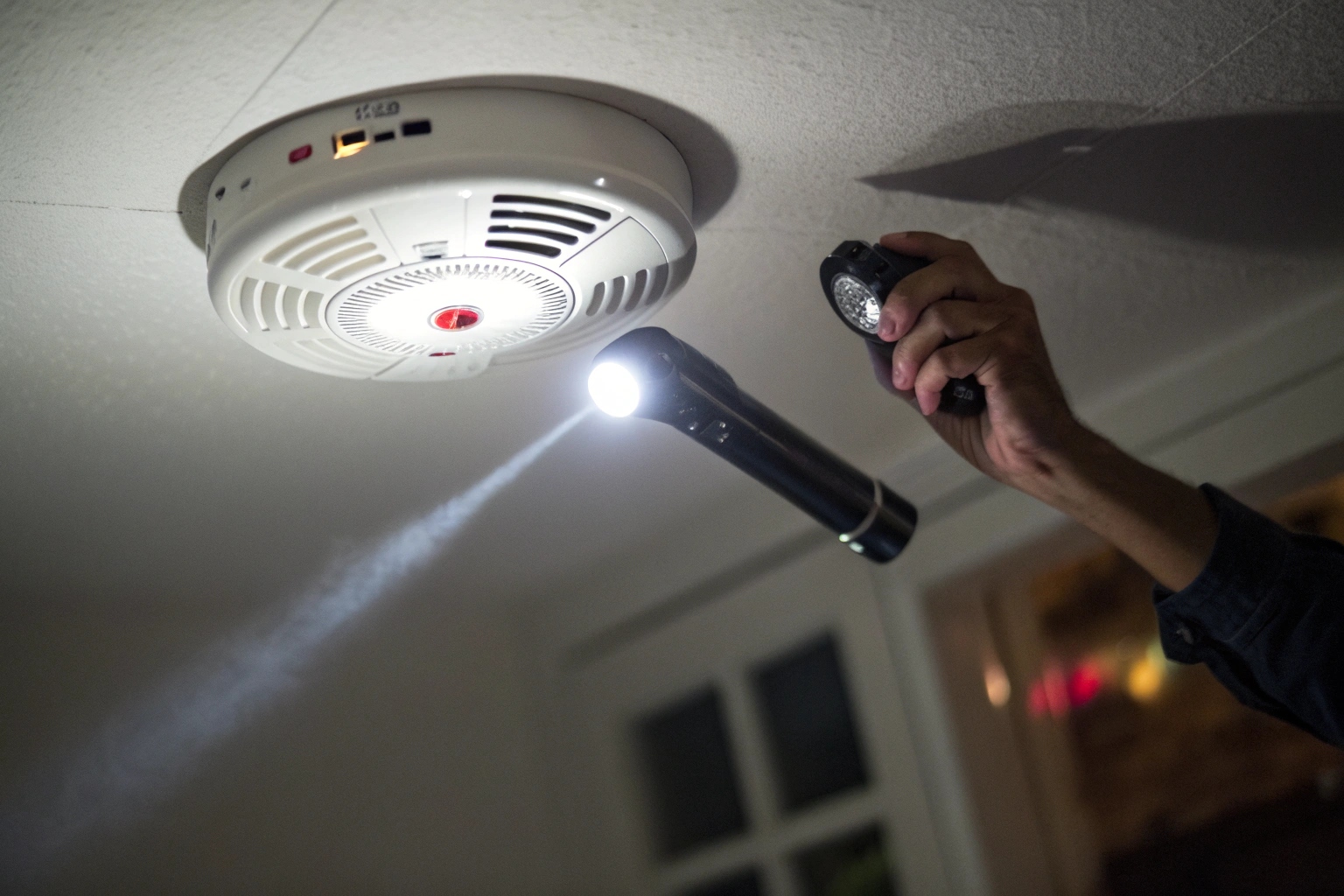 A close-up shot of a hand using a flashlight to reveal the glint of a tiny camera lens hidden in a smoke detector